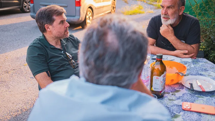 Tre uomini conversano attorno a un tavolo all’aperto.